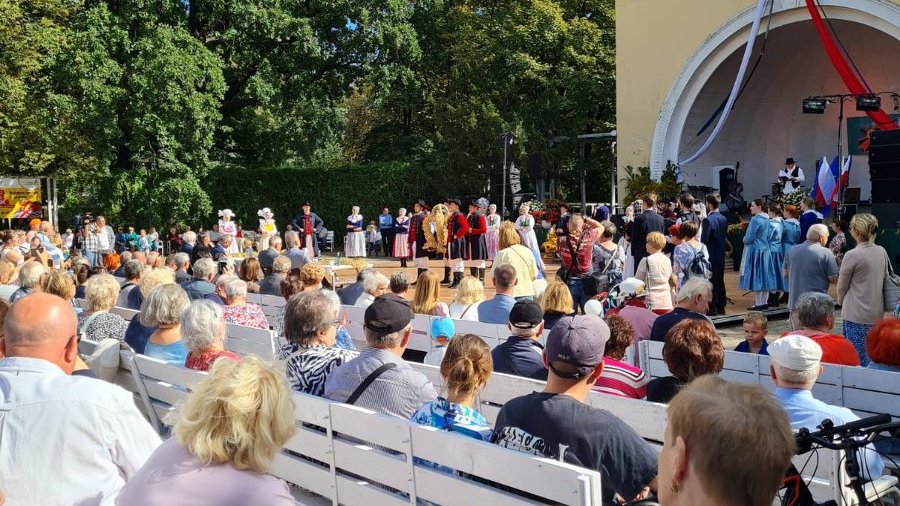 Na pierwszym planie grupa osób spogląda na scene, na której widać grupę osób w ludowych strojach.