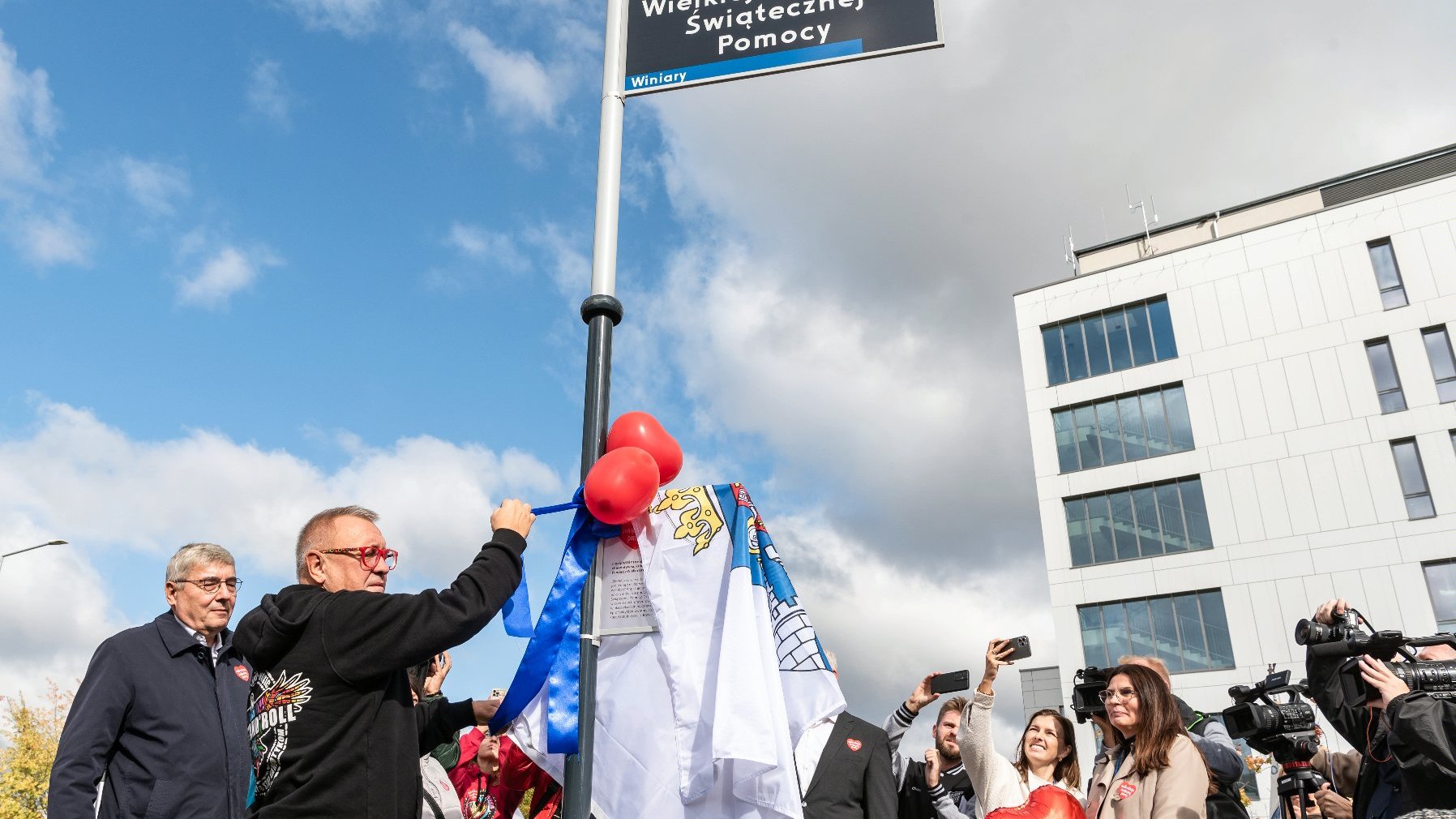 Na zdjęciu odsłonięcie tablicy z opisem skweru, Jurek Owsiak ciągnie za wstążkę, wokół samorządowcy i wolontariusze