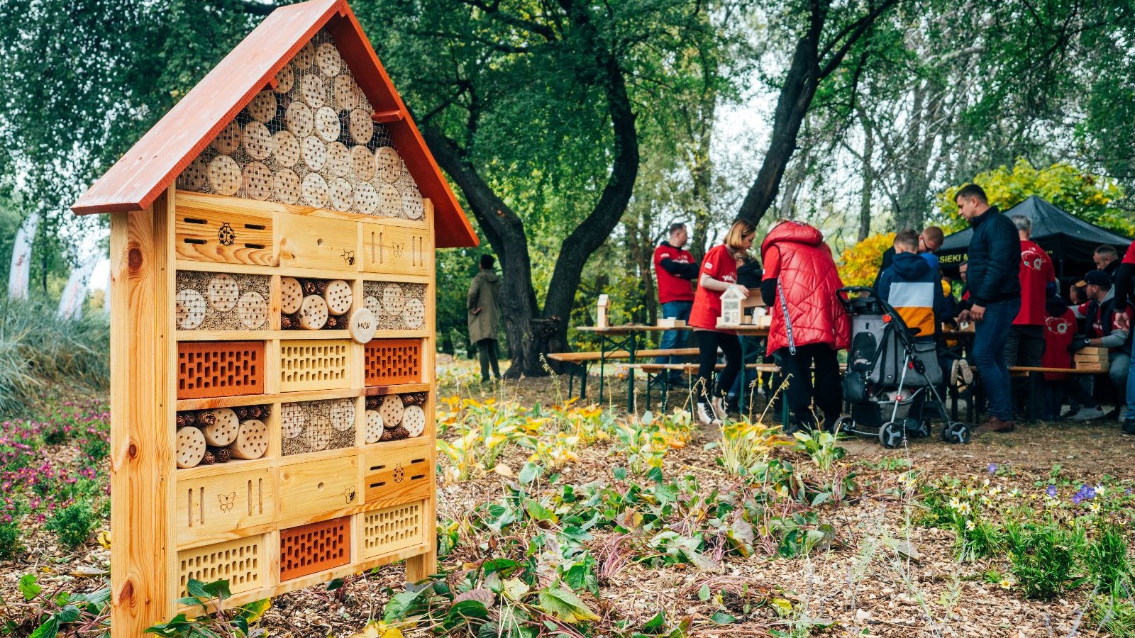 Na zdjęciu drewniany domek dla pszczół, w tle ludzie przy stolikach