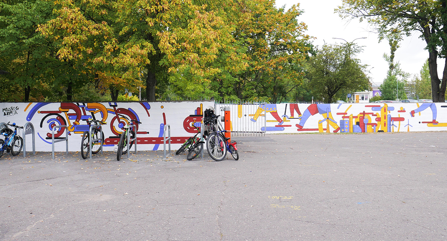 Zdjęcie przedstawia mural wykonany na ogrodzeniu szkoły, przed nim stoją rowery - grafika rozmowy