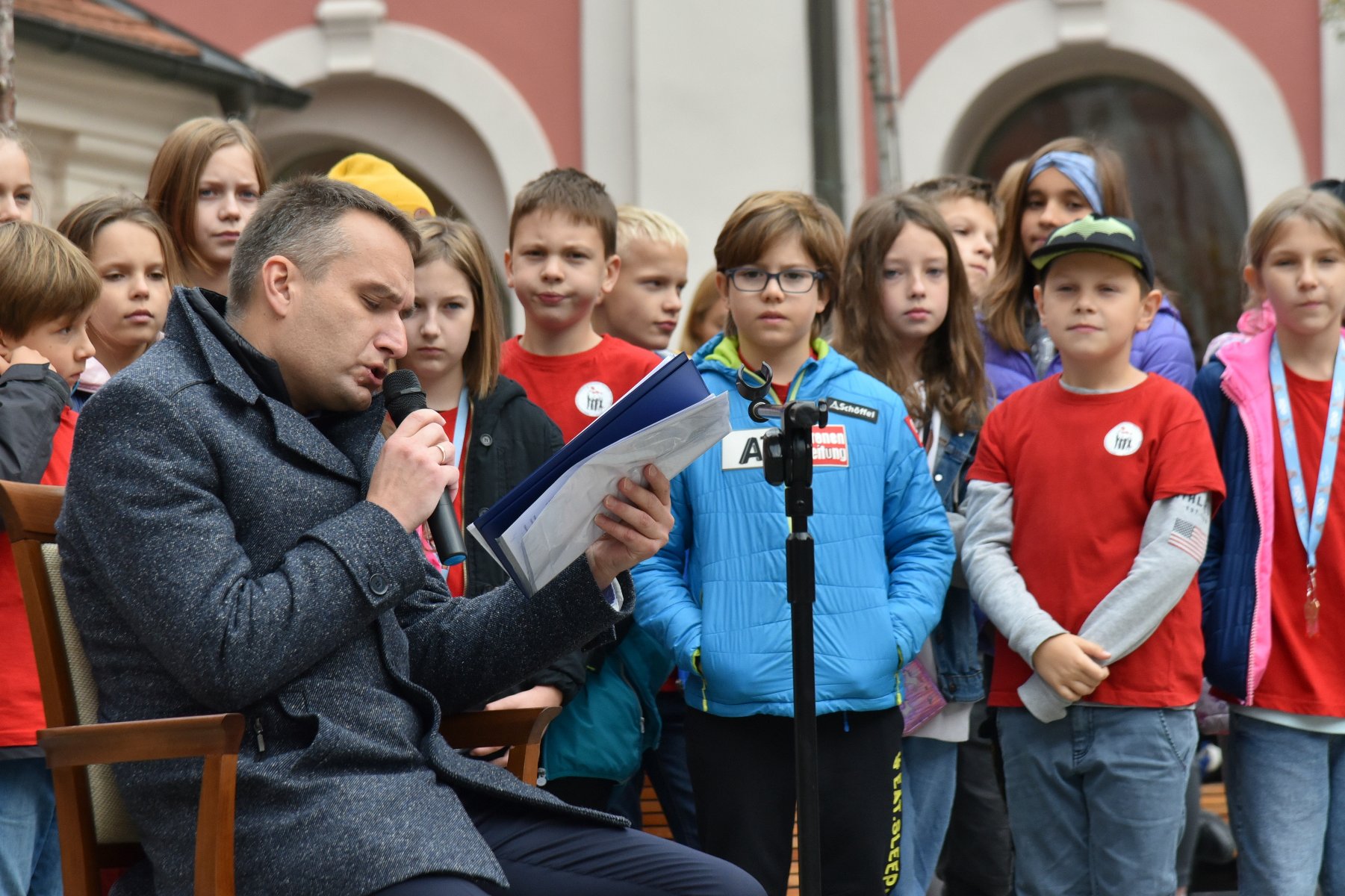 Na zdjęciu Mariusz Wiśniewski, zastępca prezydenta Poznania, czytający tekst, za nim dzieci - grafika rozmowy