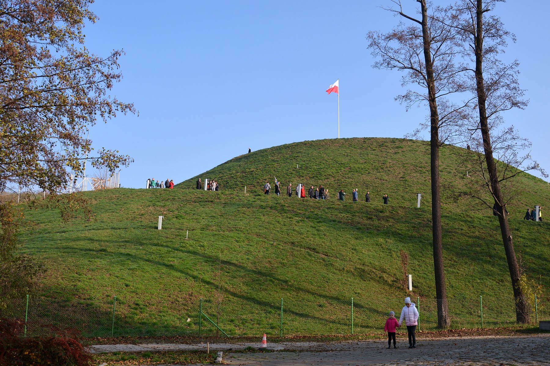 Zdjęcie przedstawia Kopiec Wolności w Poznaniu oraz wchodzących na niego ludzi. Na szczycie widąć flagę Polski. - grafika rozmowy