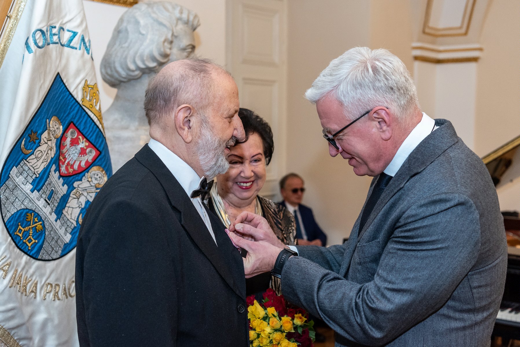 Na zdjęciu Jacek Jaśkowiak, prezydent Poznania, przypina medal mężczyźnie - grafika rozmowy