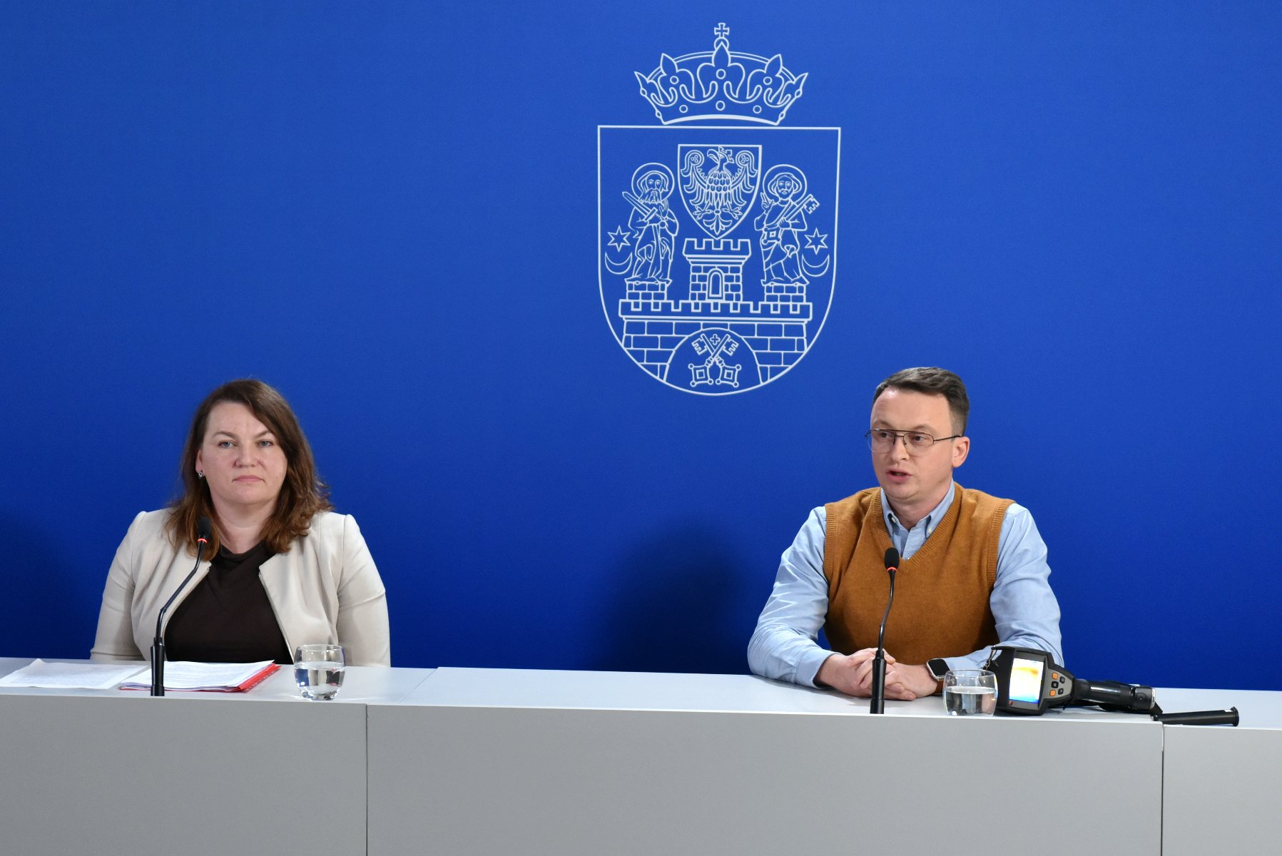 Dwie osoby za stołem konferencyjnym, mężczyzna i kobieta, za nimi granatowa ścianka z herbem miasta - grafika rozmowy