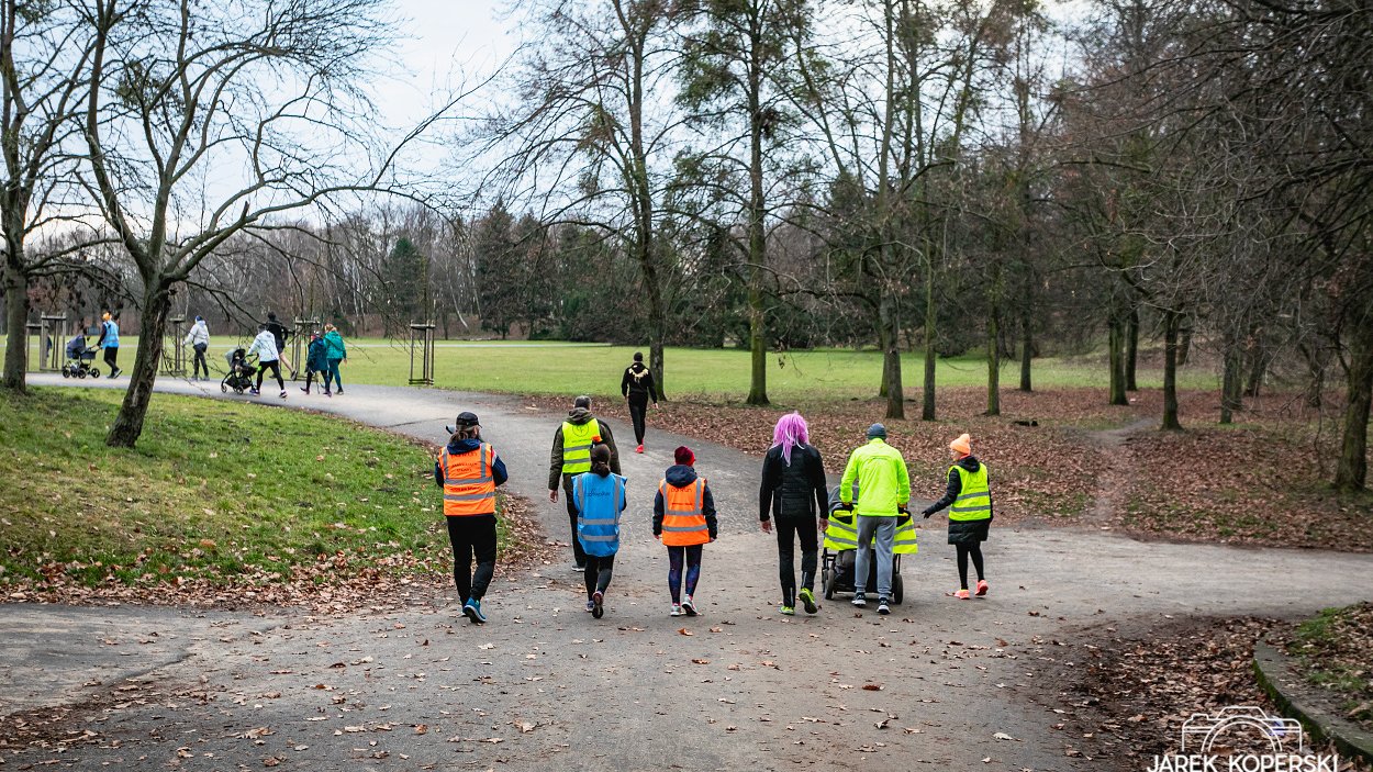 Na zdjęciu grupa ludzi w kmizelkach idąca przez park, niektórzy biegną