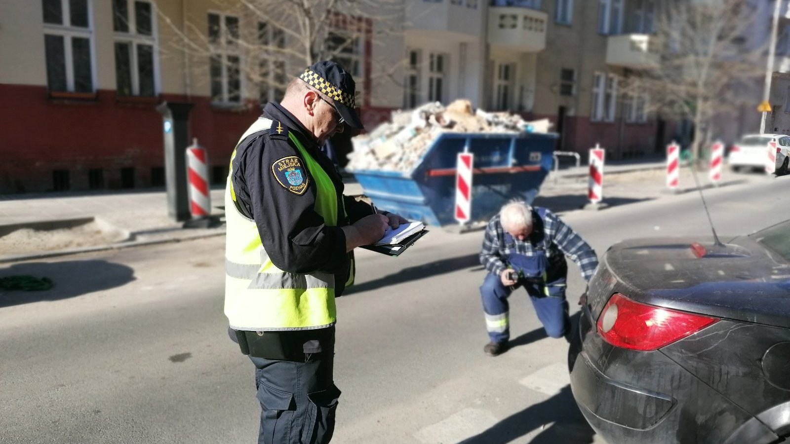 Na zdjęciu strażnik miejski zapisujący dane samochodu, w tle mężczyzna w roboczym ubraniu