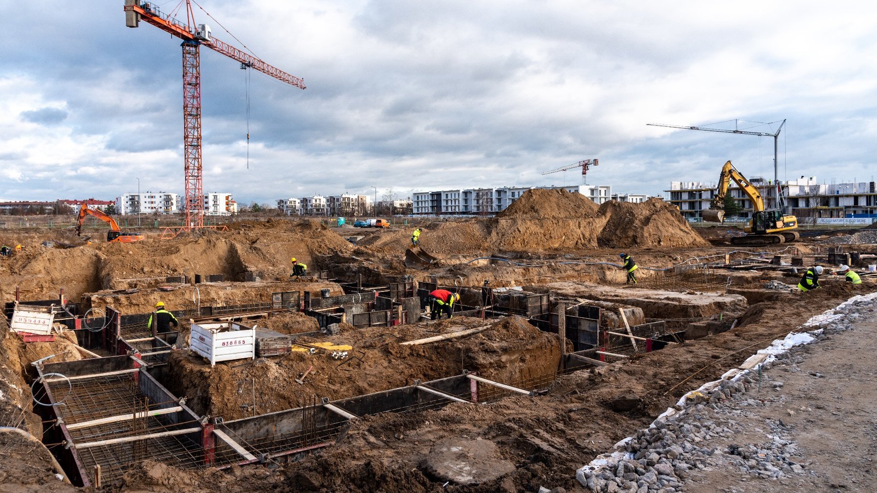 Plac budowy. Na pierwszy planie widać powstające fundamenty budynków, w tle hałdy piasku, koparka, żuraw. Po plau budowy chodzą robotnicy ubrani w kaski i kamizelki odblaskowe.