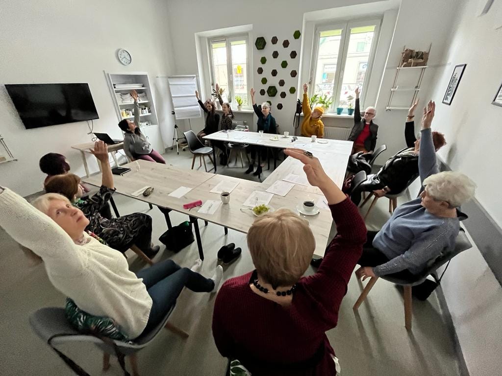 Na zdjęciu grupa ludzi przy stole, każdy z nich ma podniesioną w górę rękę - grafika rozmowy