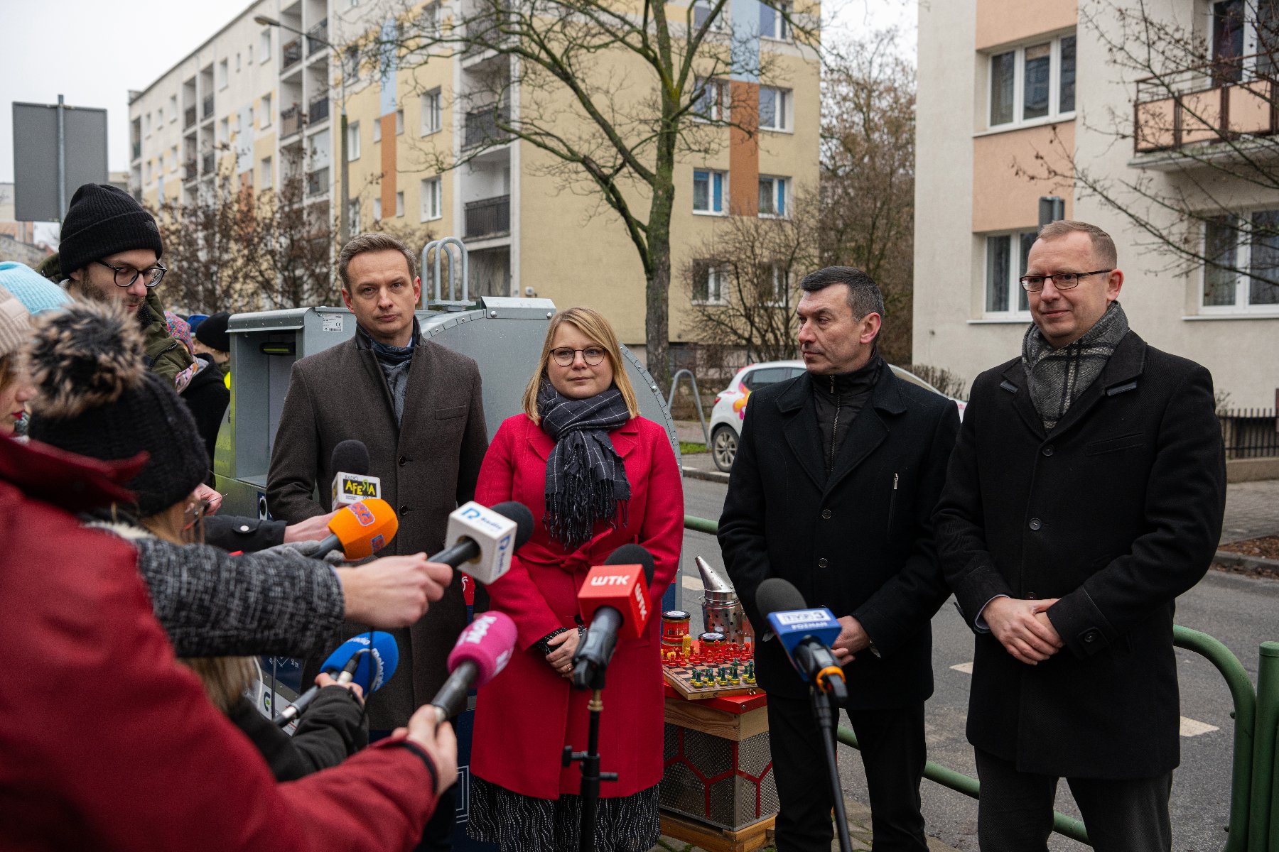 Na zdjęciu trzech mężczyzn i kobieta, obok nich dziennikarze z mikrofonami, w tle pojemnik na odpady - grafika rozmowy