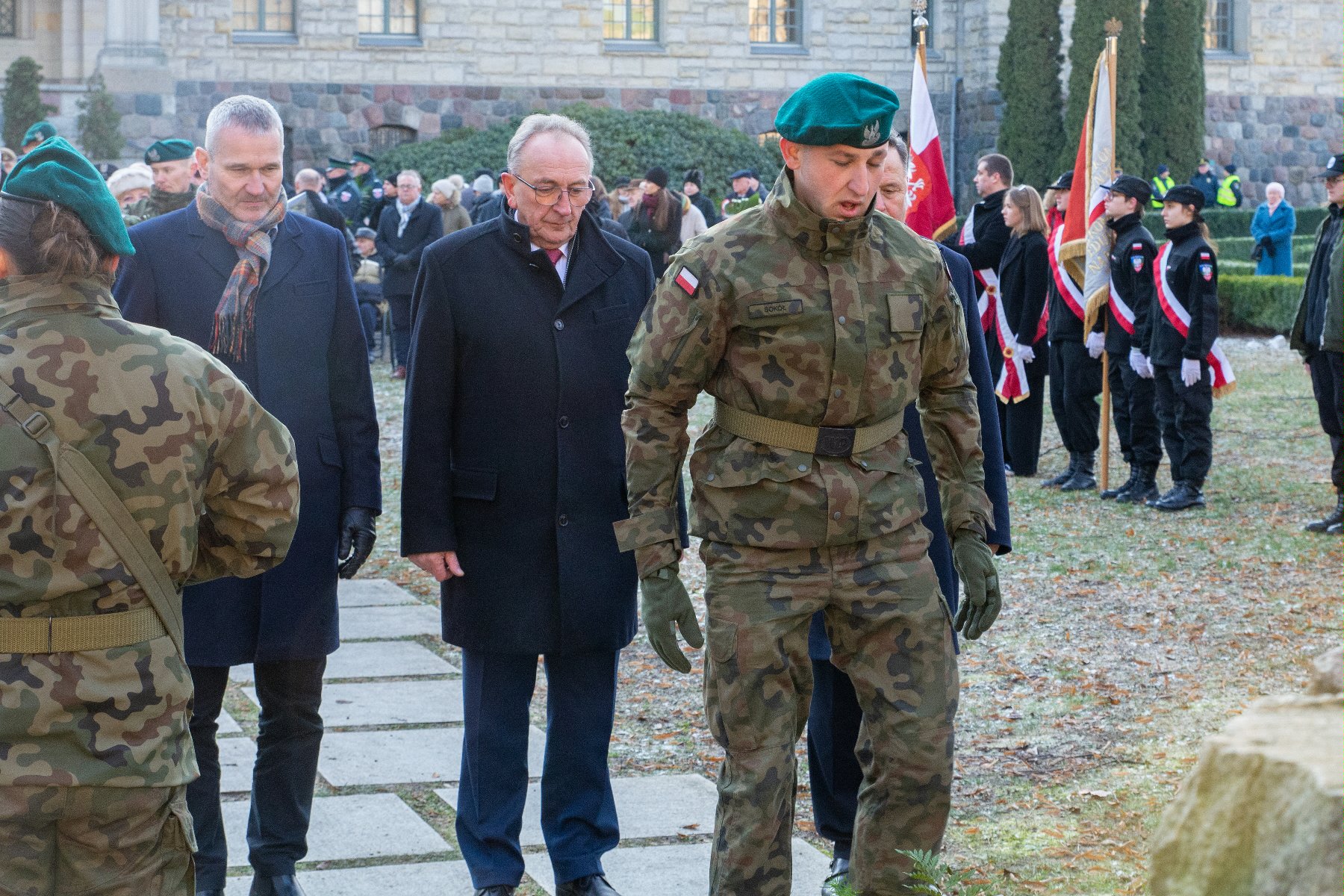 Na zdjęciu mężczyźni składający kwiaty, na pierwszym planie żołnierz - grafika rozmowy