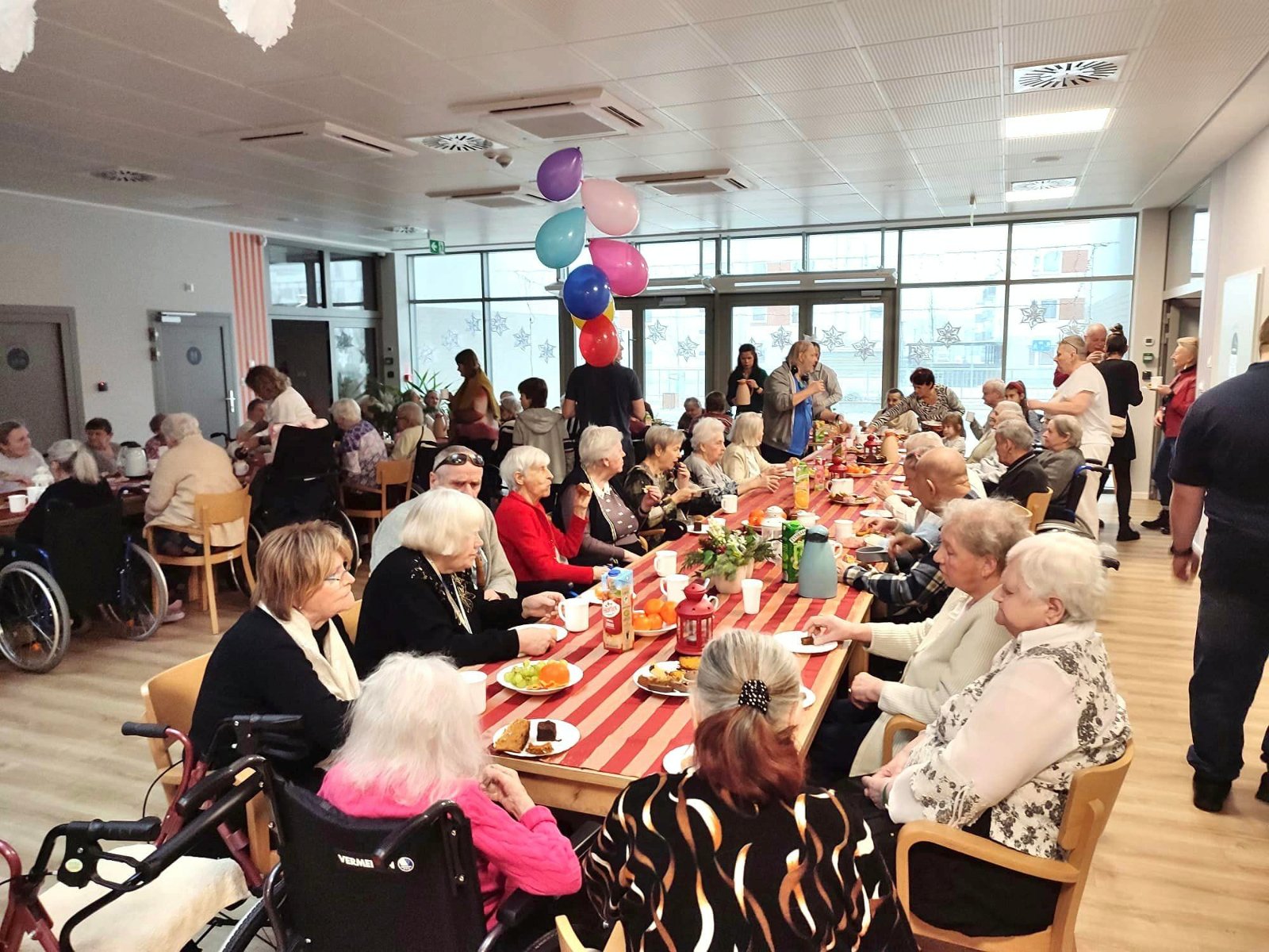 Na zdjęciu duża grupa seniorów i seniorek siedzących przy stołach - grafika rozmowy