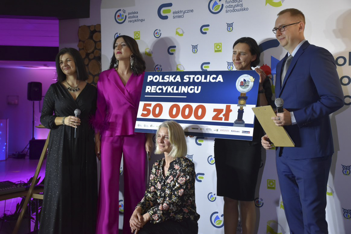 Na zdjęciu piątka ludzi w eleganckich strojach trzymająca bon na 50 000 złotych - grafika rozmowy