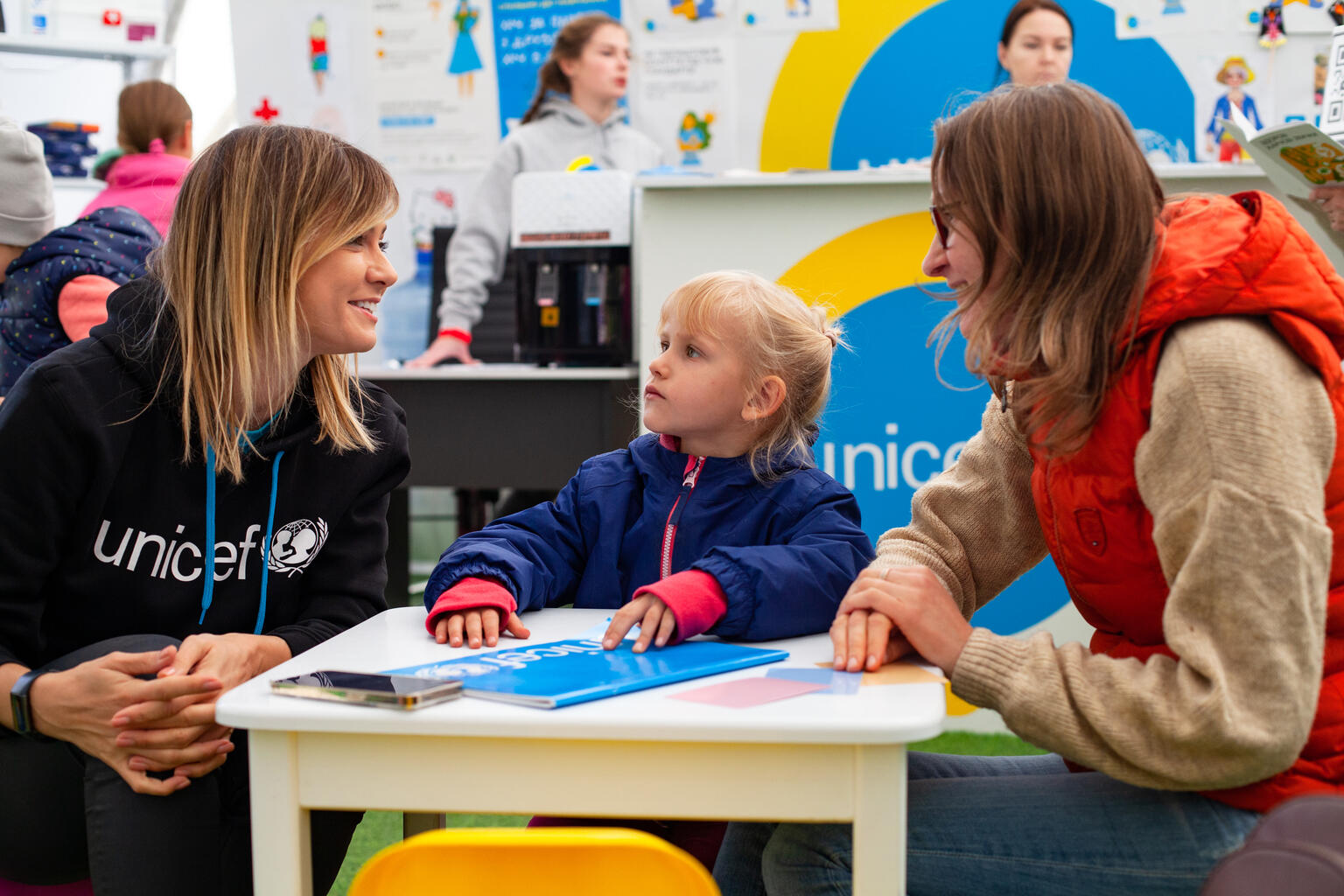 Na zdjęciu dwie kobiety i dziewczynka przy małym stole, jedna z kobiet ma bluzę z logotypem UNICEF - grafika rozmowy