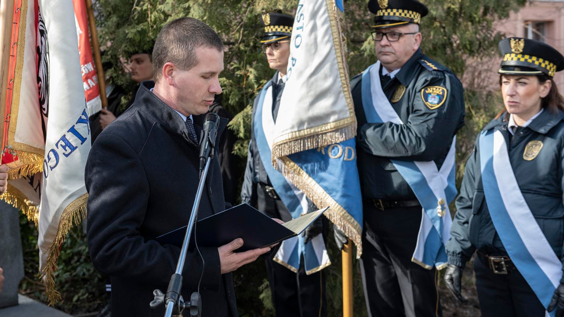 Mężczyzna przy mikrofonie, czyta coś z kartki, w tle poczet sztandarowy