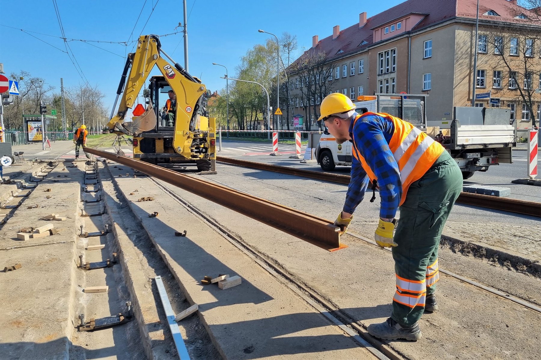 Zdjęcie przedstawia pracowników służb torowych układającyh torowisko. W tle koparka. - grafika rozmowy