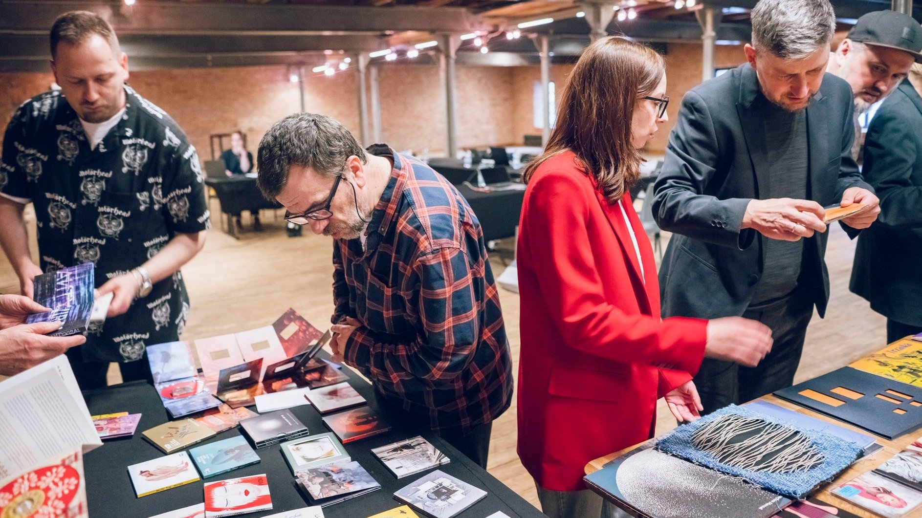 Zdjęcie przedstawia grupę osób, które nachylają się nad stołem, na którym leżą okładki płyt CD oraz winylowych.