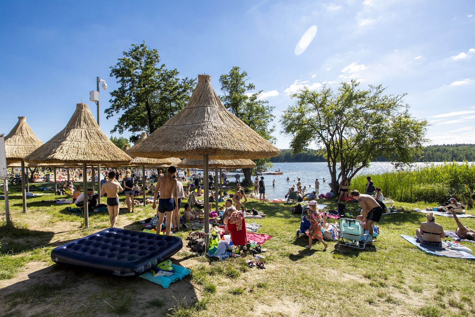 Zdjęcie przedstawia kąpielisko Strzeszynek. Na pierwszym planie widać grupę ludzi odpoczywającą na ręcznikach. W tle jezioro. - grafika rozmowy