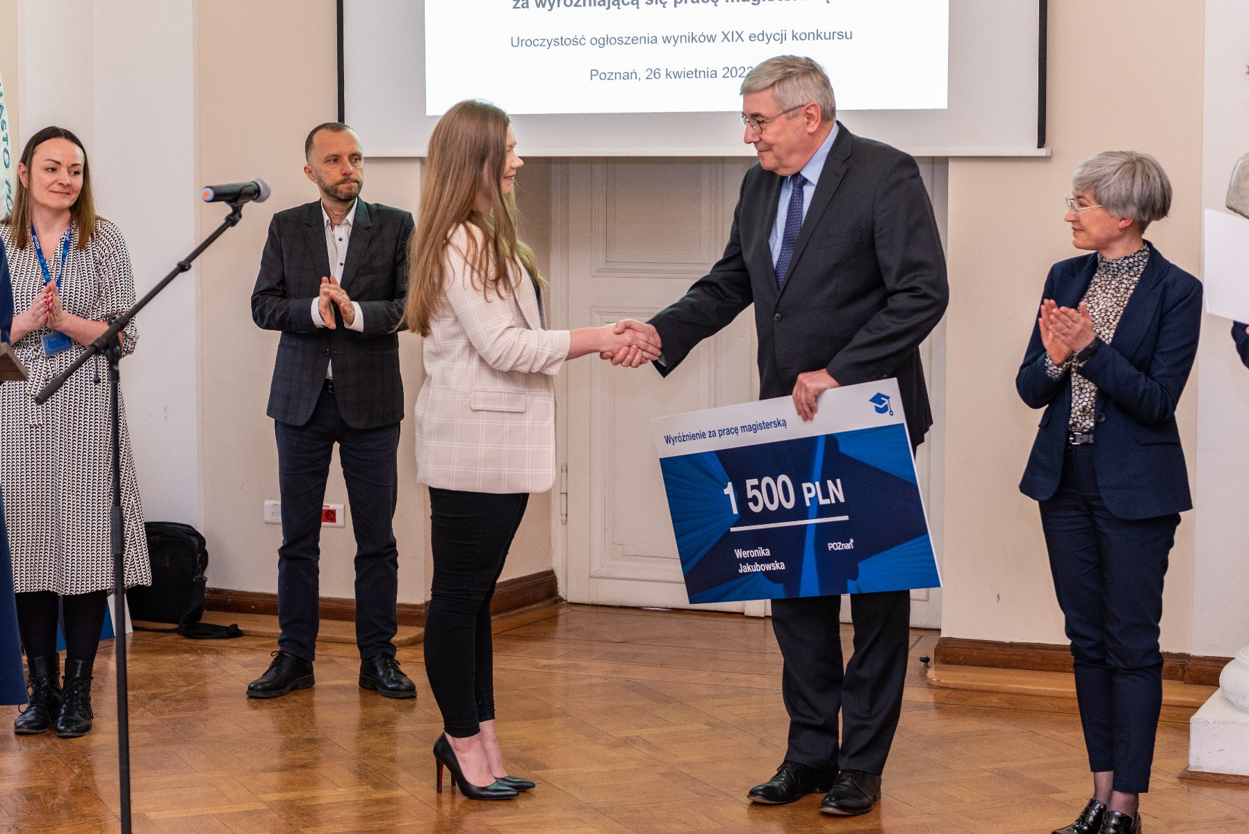 Na zdjęciu przedwodniczący rady miasta wręcza czek jednej z laureatek, w tle stoi trójka ludzi - grafika rozmowy