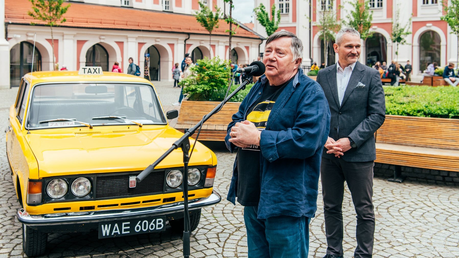 Galeria zdjęć z konferencji na dziedzińcu urzędu miasta, na zdjęciach żółta taksówka z serialu "Zmiennicy", aktor oraz zastępca prezydenta Poznania