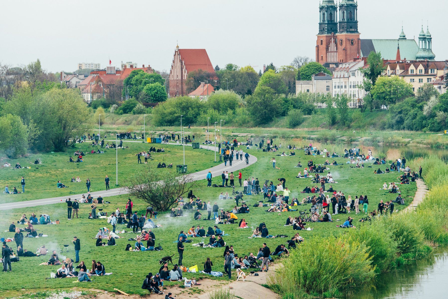 Zdjęcie przedstawia grupę ludzi odpoczywających nad Wartą. W tle katedra. - grafika rozmowy