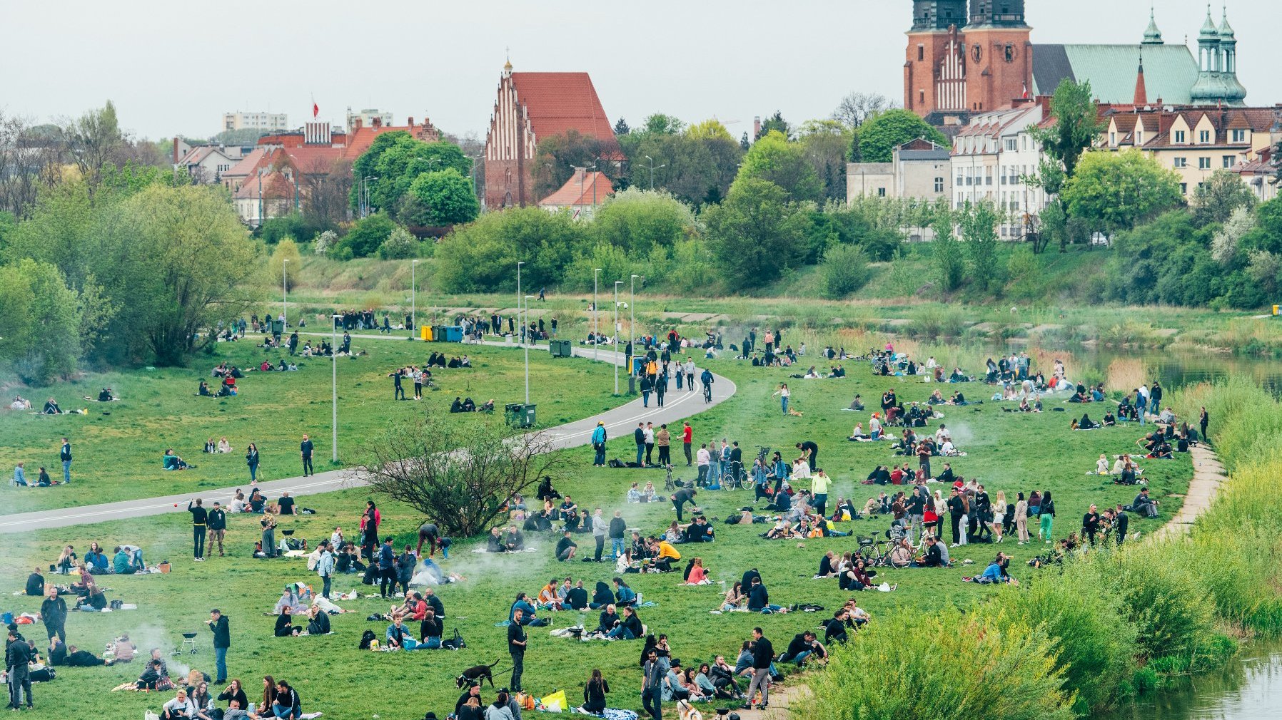 Zdjęcie przedstawia grupę ludzi odpoczywających nad Wartą. W tle katedra.