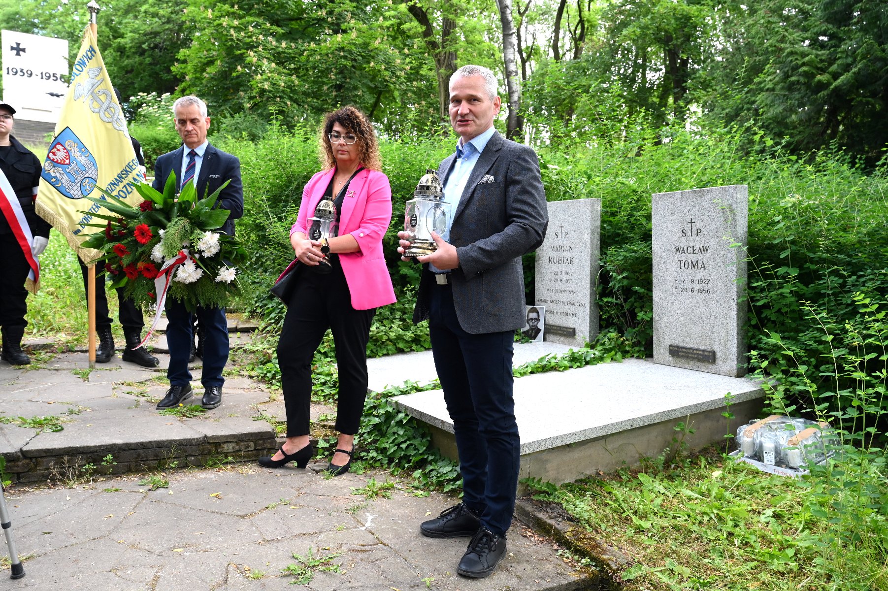 Na zdjęciu trójka ludzi stojąca przeg grobami, mężczyźni trzymają wieniec i znicz, a kobieta znicz - grafika rozmowy