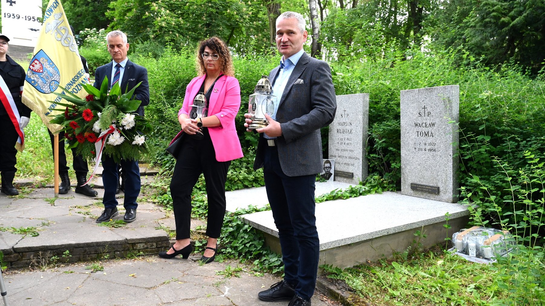 Na zdjęciu trójka ludzi stojąca przeg grobami, mężczyźni trzymają wieniec i znicz, a kobieta znicz