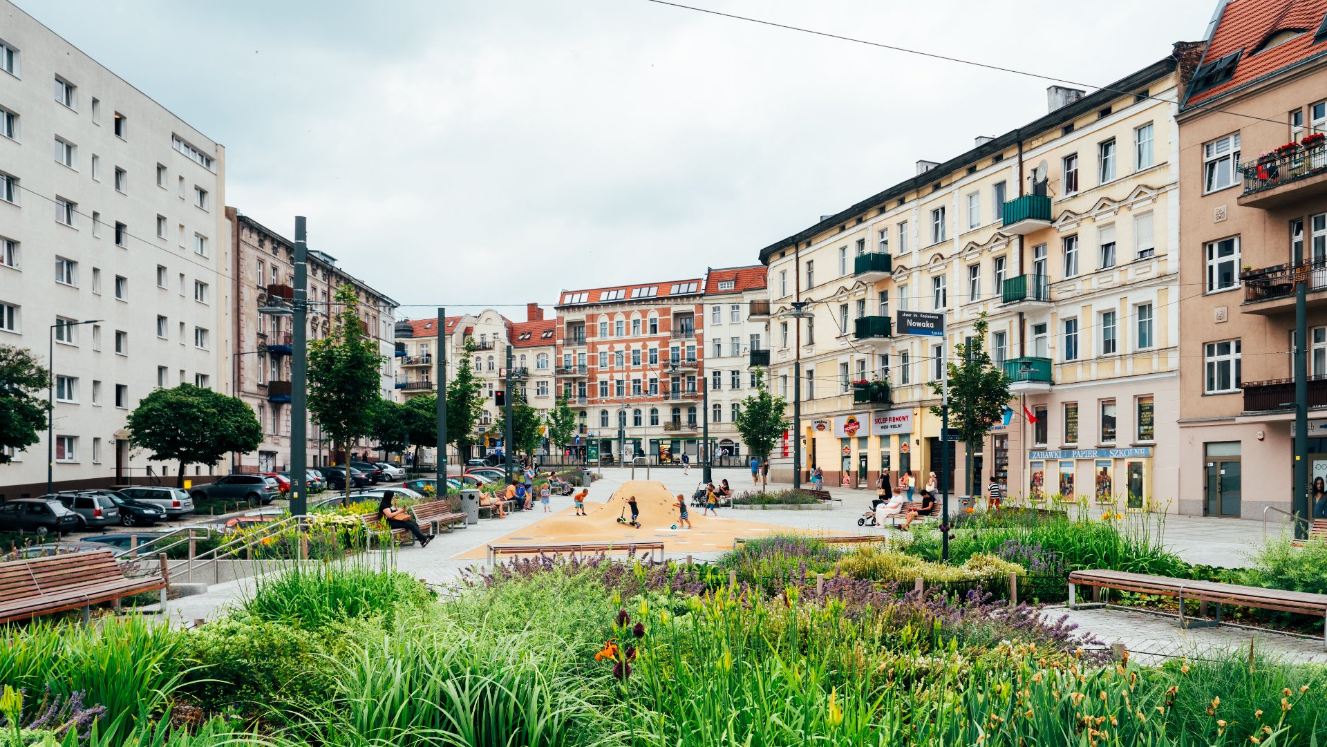 Na zdjęciu rynek Łazarski, na pierwszym planie rośliny, w tle bawiące się dzieci - grafika rozmowy