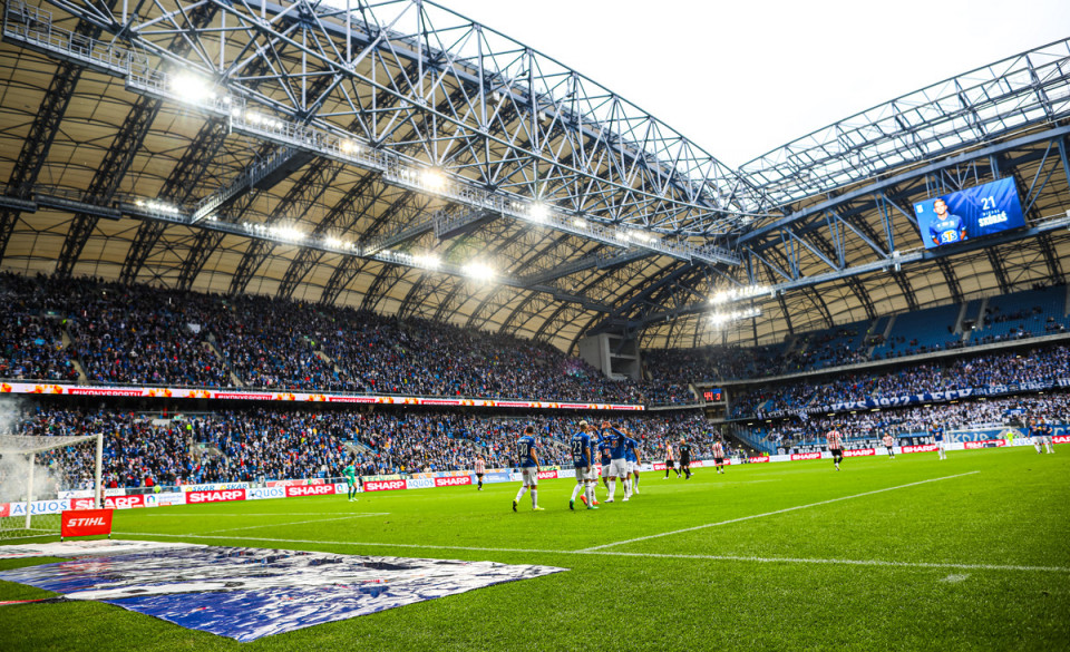 Zdjęcie przedstawia stadion Lecha. Na pierwszym planie widać piłkarzy na murawie w tle trybuny. - grafika rozmowy