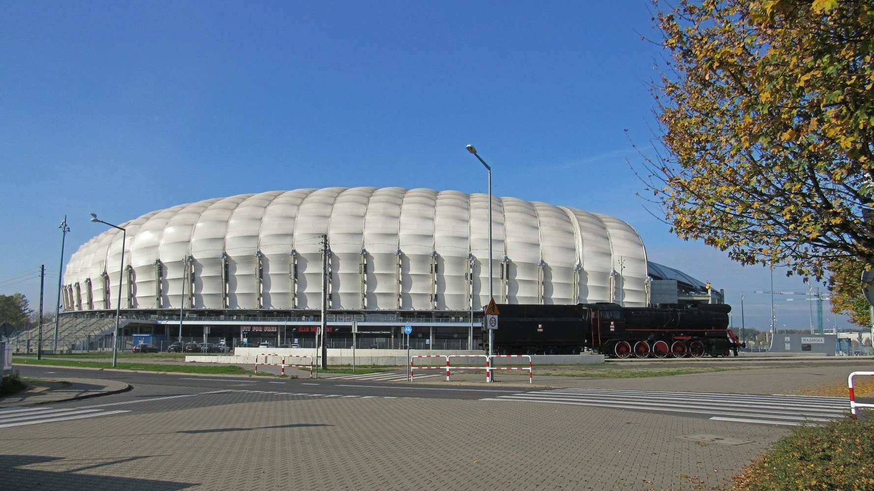 Zdjęcie przedstawia białą kopułę stadionu przy ul. Bułgarskiej. Przed nim stoi parowóz.