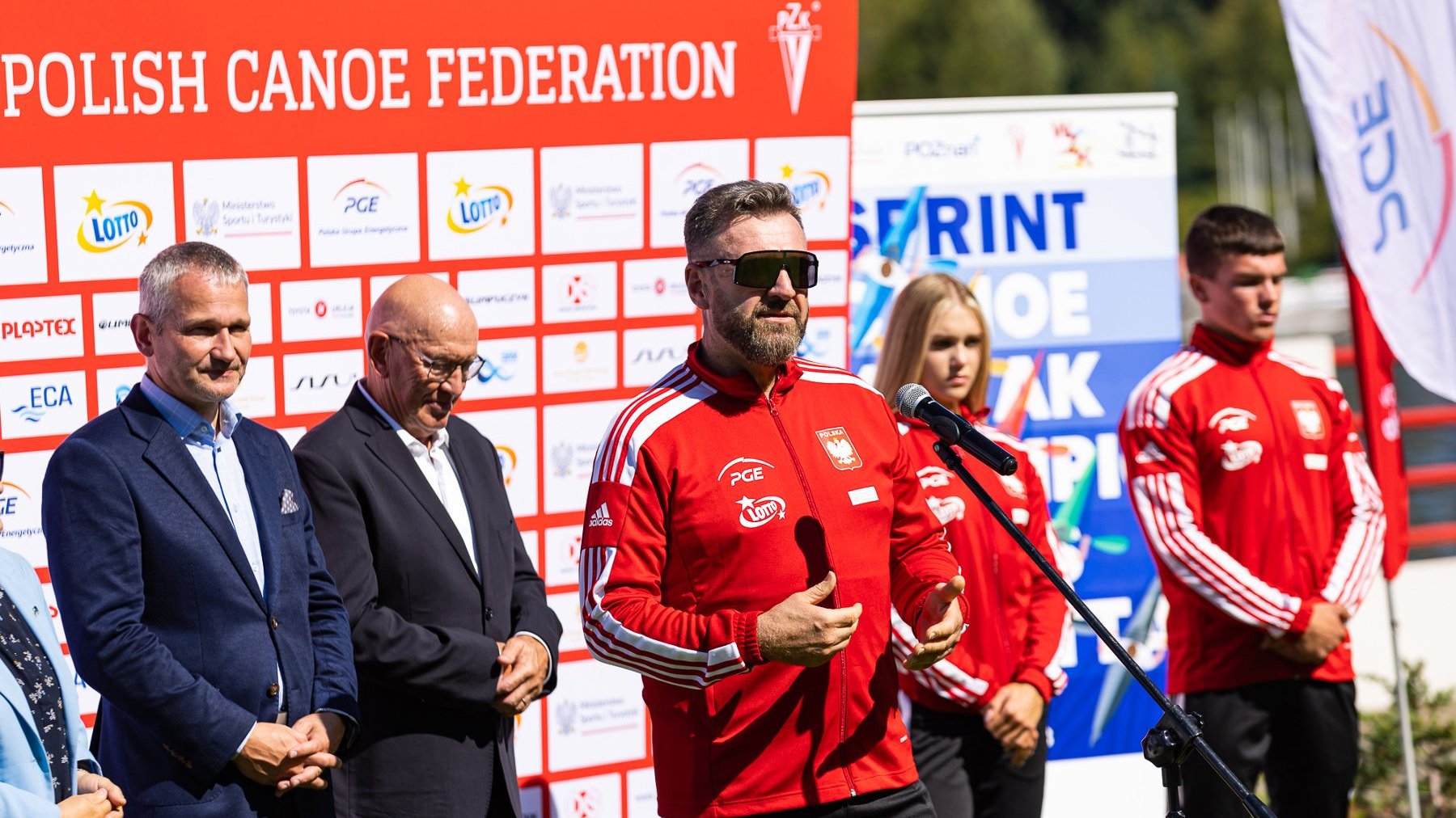 Na zdjęciu konferencja prasowa, przy mikrofonie mężczyzna w sportowej bluzie