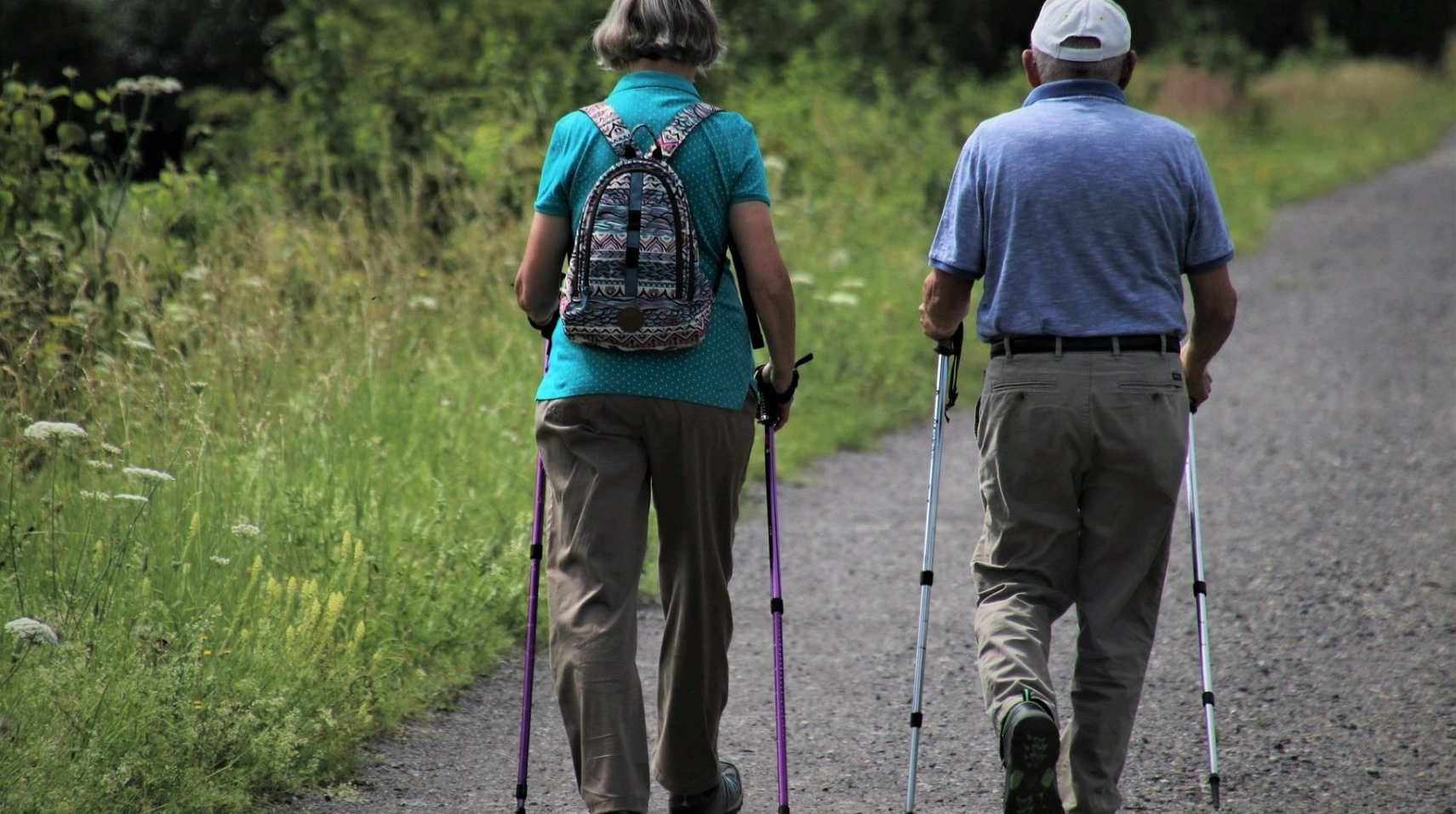 Na zdjęciu dwoje seniorów z kijkami do nordic walkingu, tyłem do obiektywu - grafika rozmowy