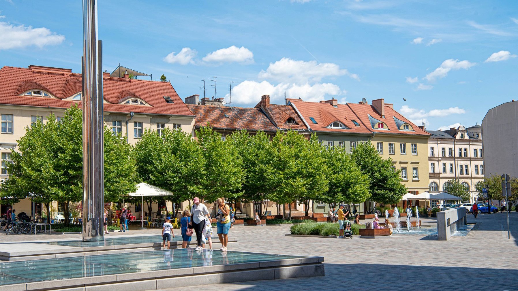 Zdjęcie przedstawia plac Kolegiacki. Widok na "zegar czasu", kaminienice i platany