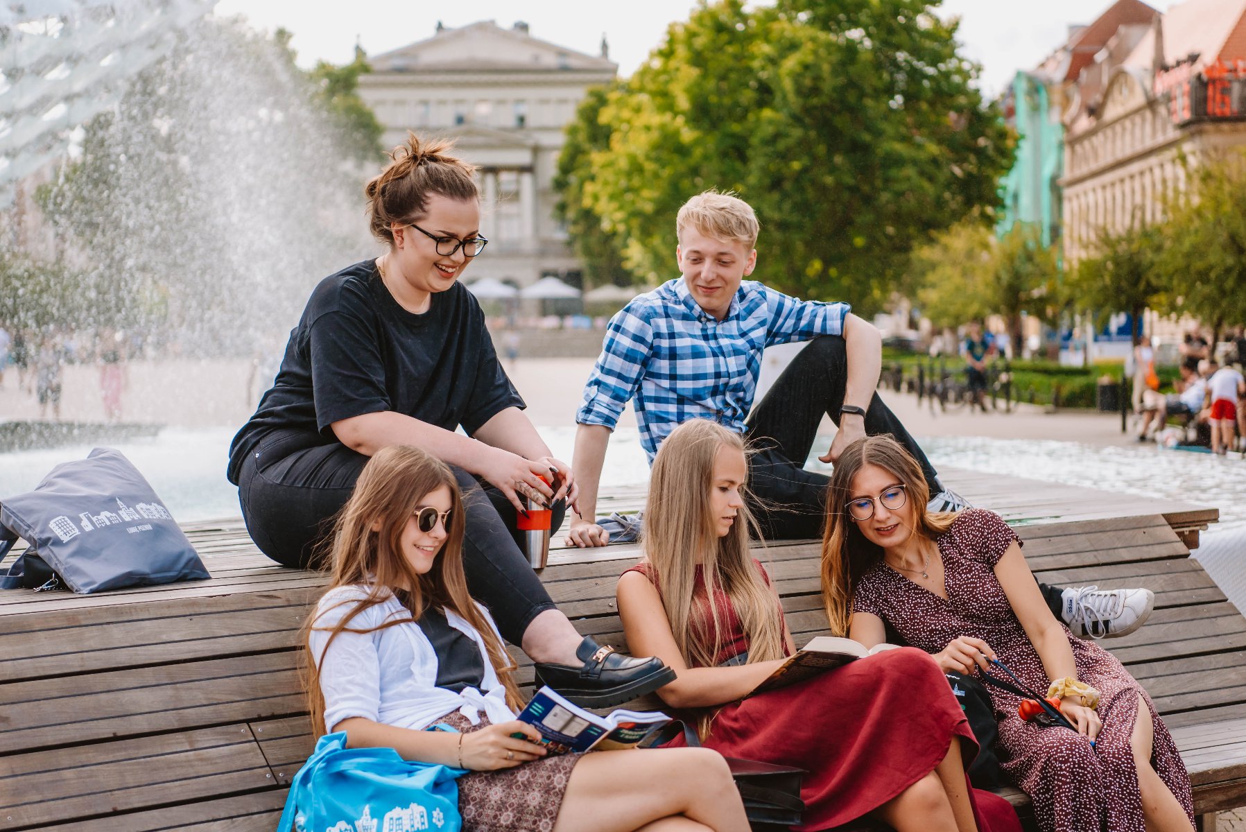 Na zdjęciu grupa młodych ludzi siedzi przy fontannie, część z nich ma w rękach książki - grafika rozmowy