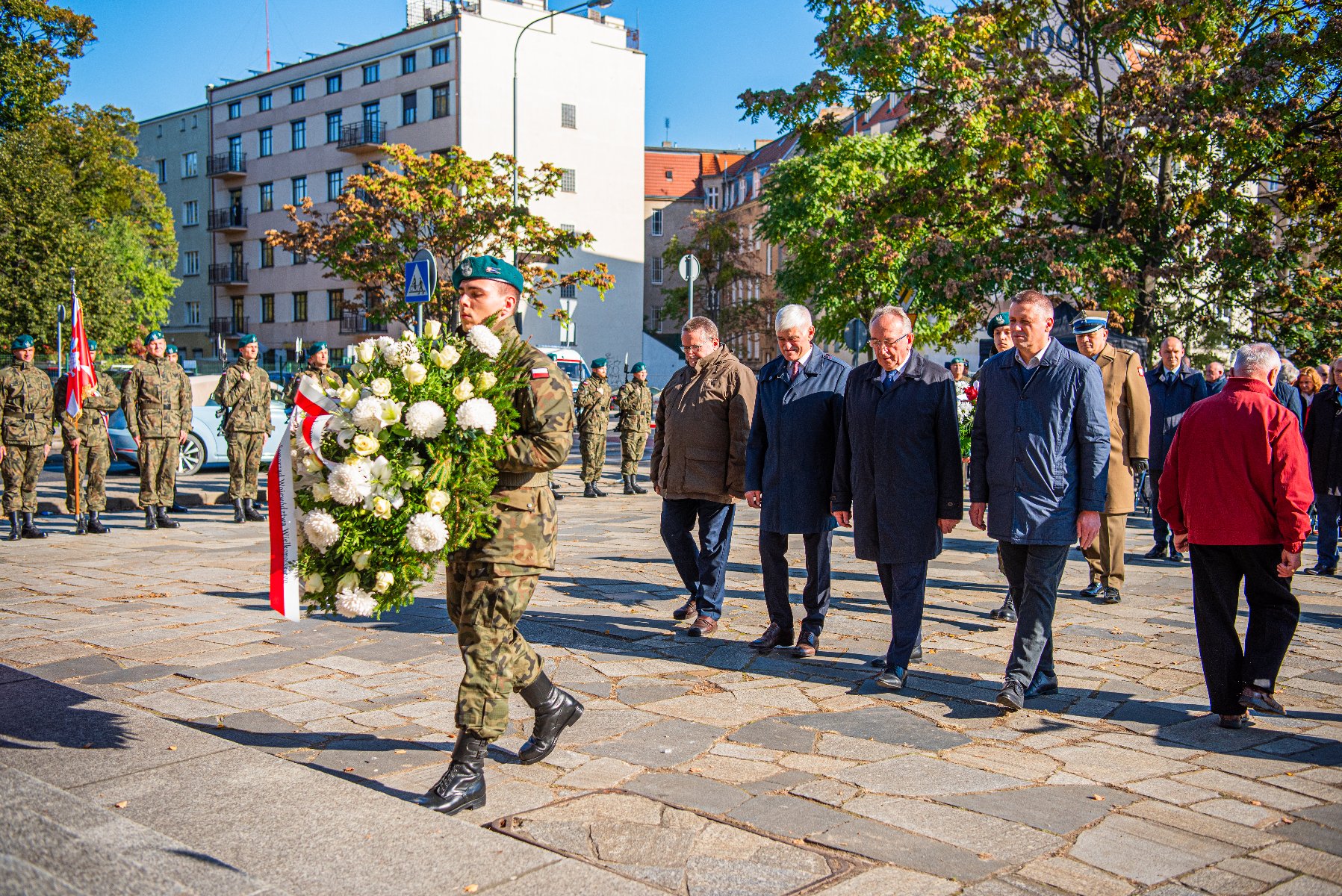 Na zdjęciu delegacja, przed nią żołnierz trzymający wiązankę - grafika rozmowy