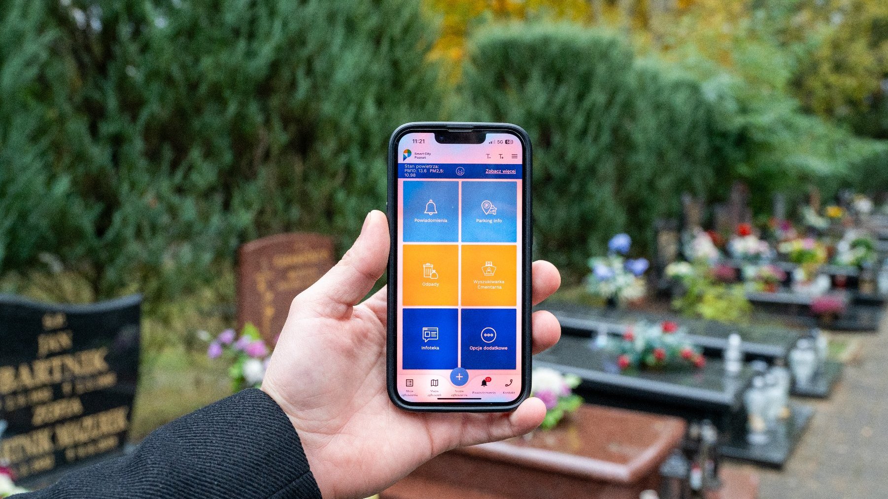 Na zdjęciu dłoń trzymająca telefon, na ekranie wyszukiwarka cmentarna, w tle groby