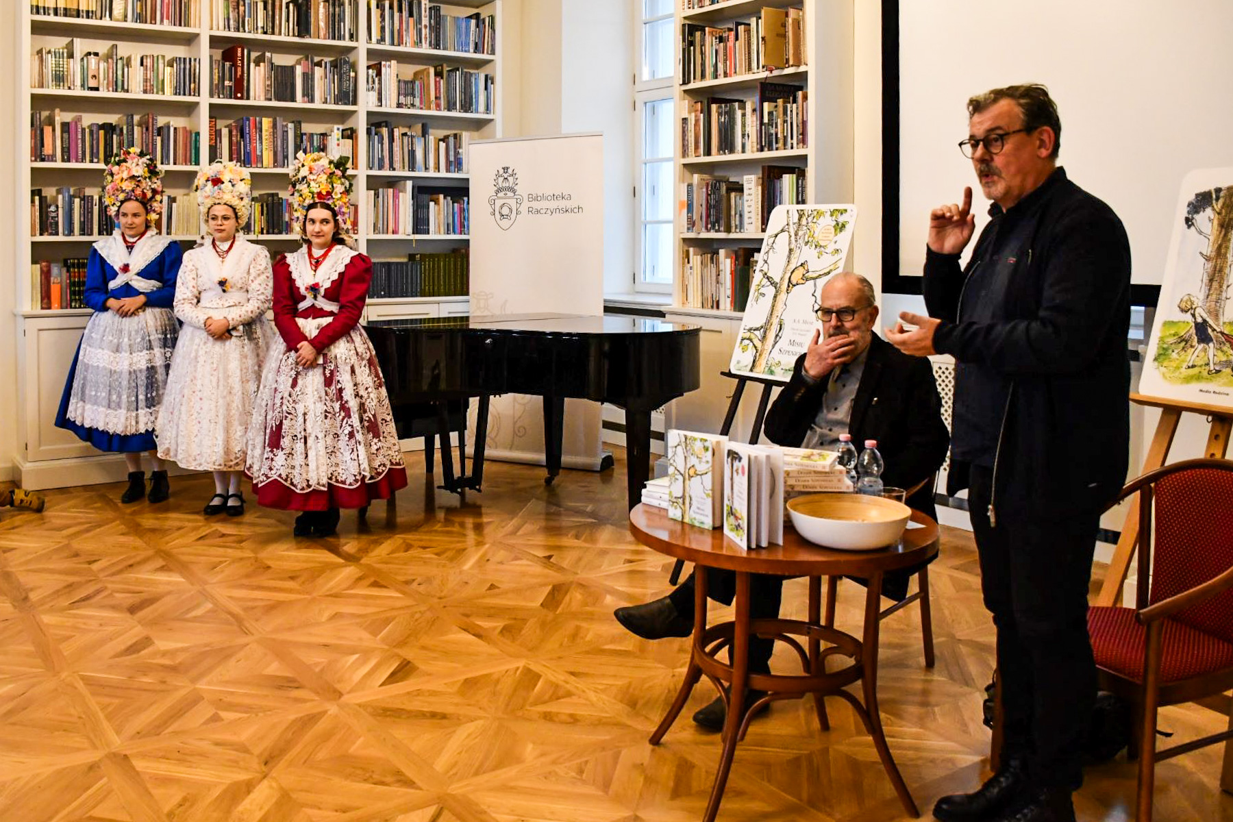 Na zdjęciu spotkanie autorskie z tłumaczem, obok trzy kobiety w strojach bamberskich - grafika rozmowy