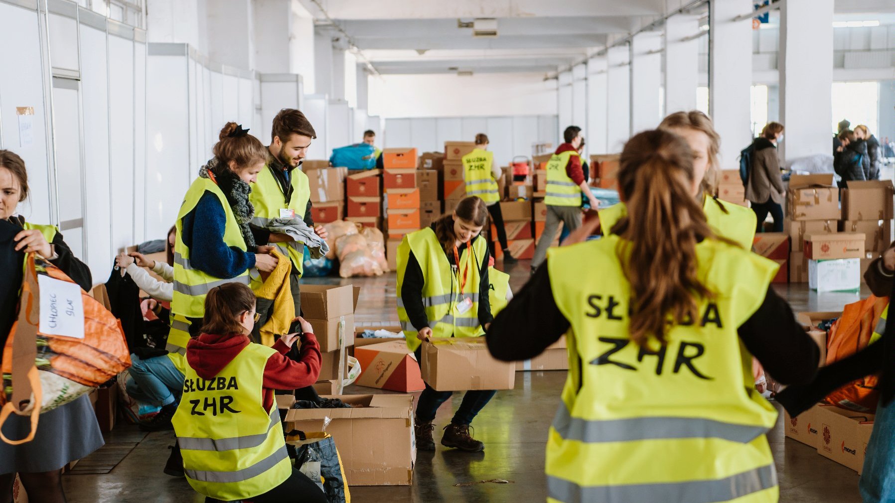 Zdjęcie przedstawia grupę osób w odblaskowych kamizelkach, które przenoszą paczki z pomocą humanitarną. - grafika rozmowy