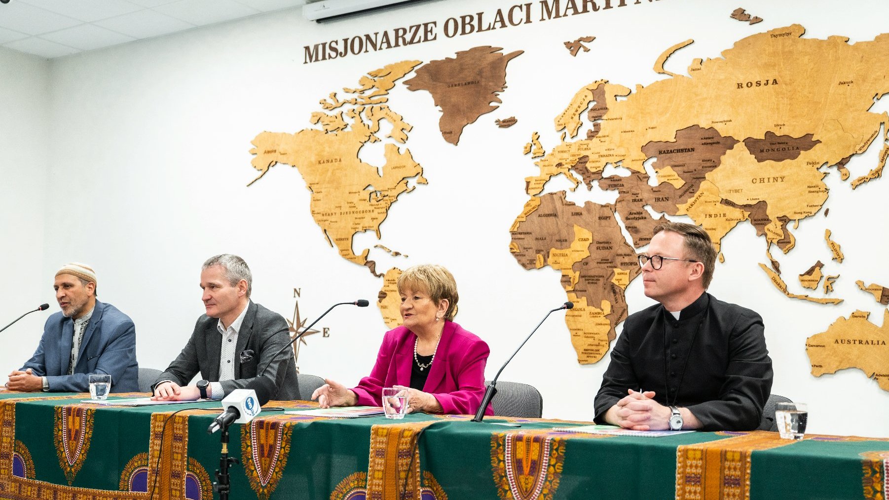 Na zdjeciu trzech mężczyzn i kobieta za stołem, w tle mapa świata na ścianie