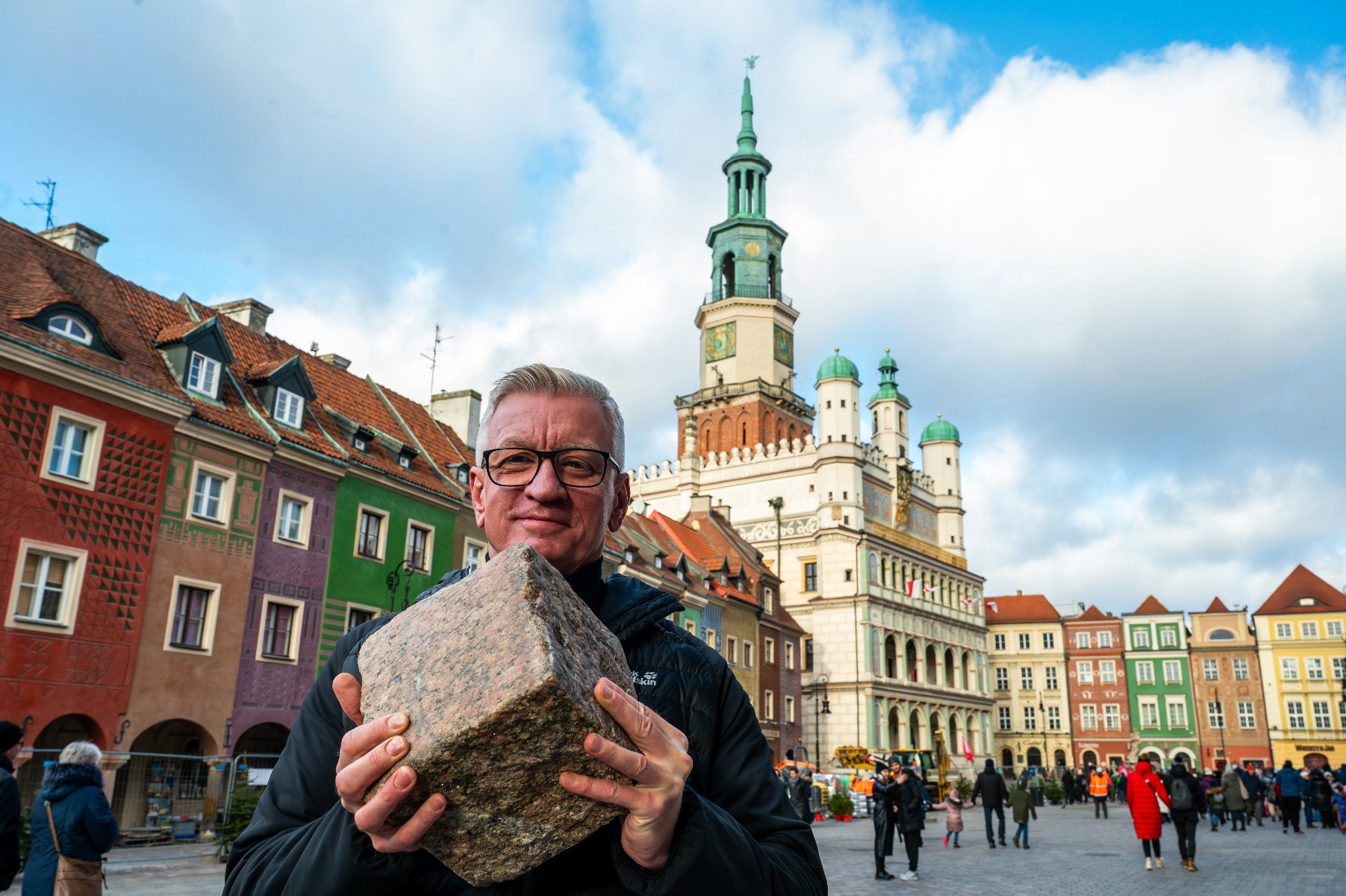 Na pierwszym planie Jacek Jaśkowiak trzymający w dłoniach kamienną kostkę. W tle ratusz i domki budnicze. - grafika rozmowy