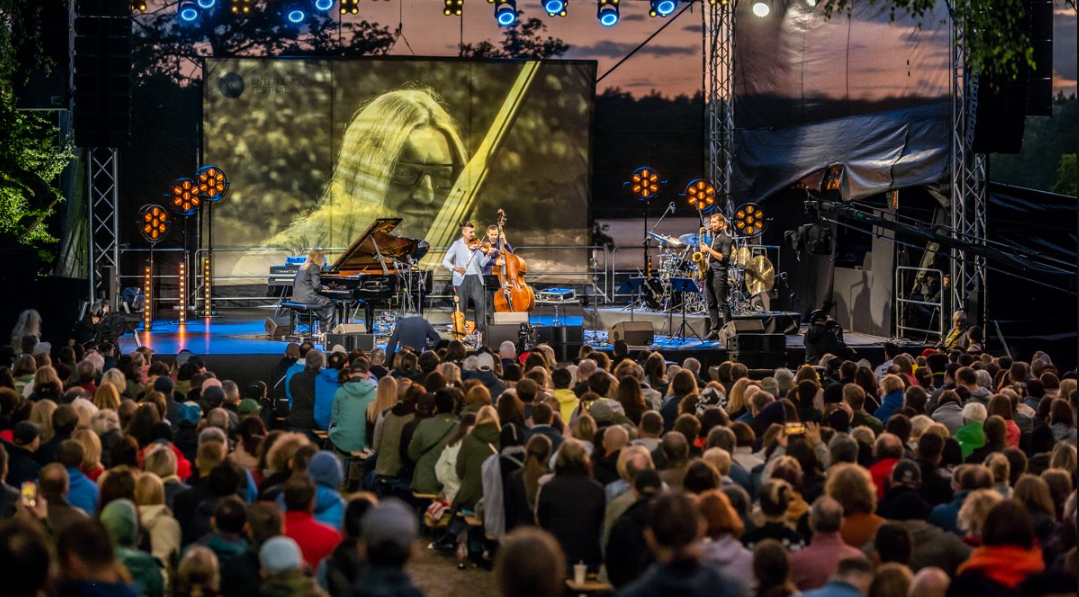 Na zdjęciu ludzie słuchający muzyki, w tle ustawiona w plenerze scena, na niej muzycy - grafika rozmowy