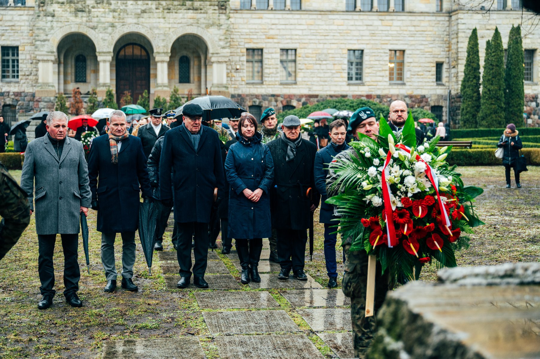 Na zdjęciu przedstawiciele władz rządowych i samorządowych stoją przed pomnikiem, przed nimi strażnik trzymający wiązankę - grafika rozmowy