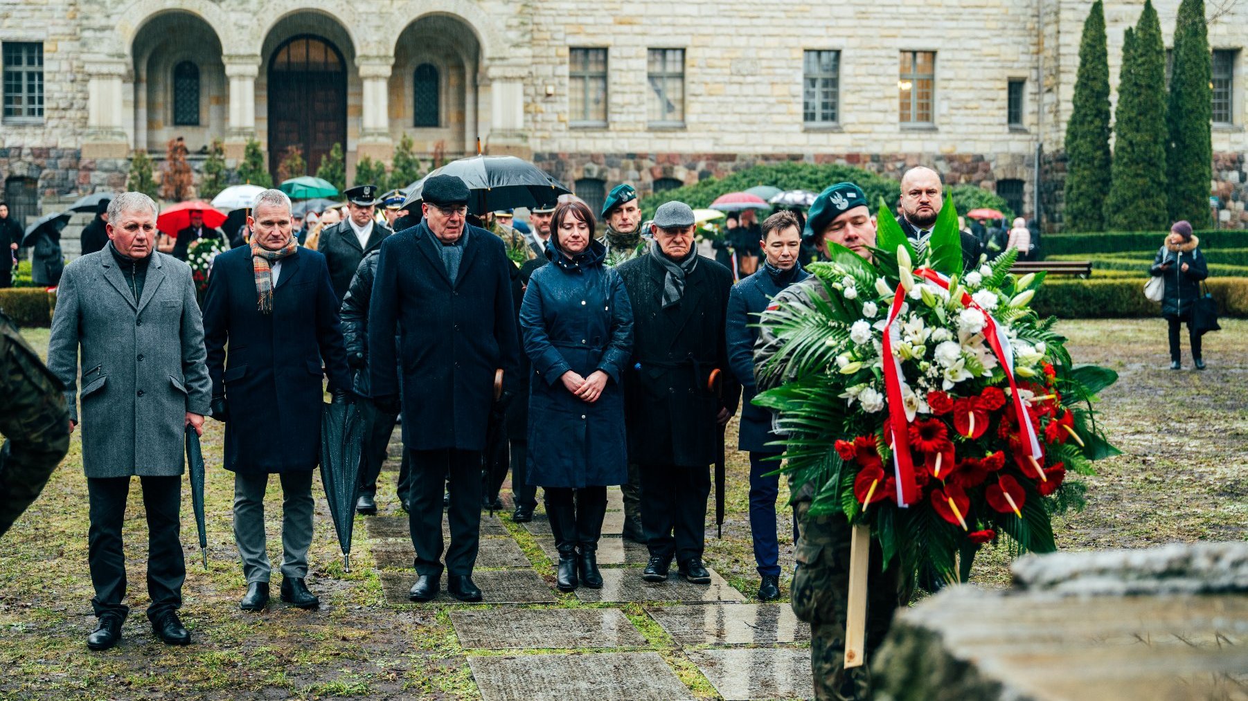 Na zdjęciu przedstawiciele władz rządowych i samorządowych stoją przed pomnikiem, przed nimi strażnik trzymający wiązankę