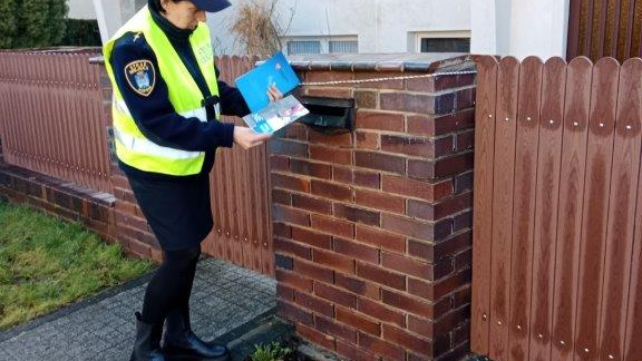 Zdjęcie przedstawia strażniczkę miejską na drodze, wrzucającą ulotkę do skrzynki na listy.