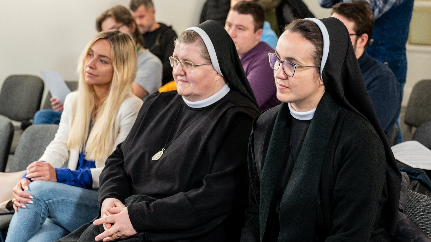 Na zdjęciu kobiety siedzące na krzesłach w sali, na pierwszym planie dwie siostry zakonne w habitach