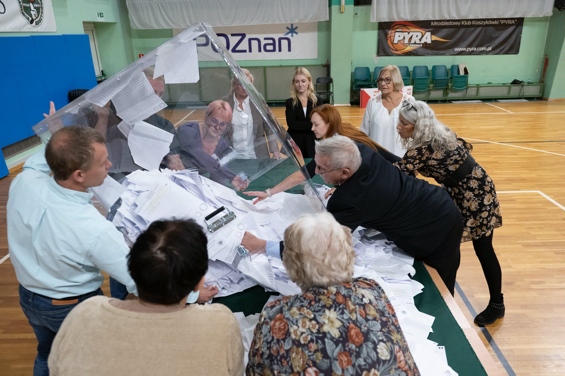 Zdjęcie przedstawia grupę osób, członków OKW, wysypujących z urny wyborczej karty do głosowania. - grafika rozmowy