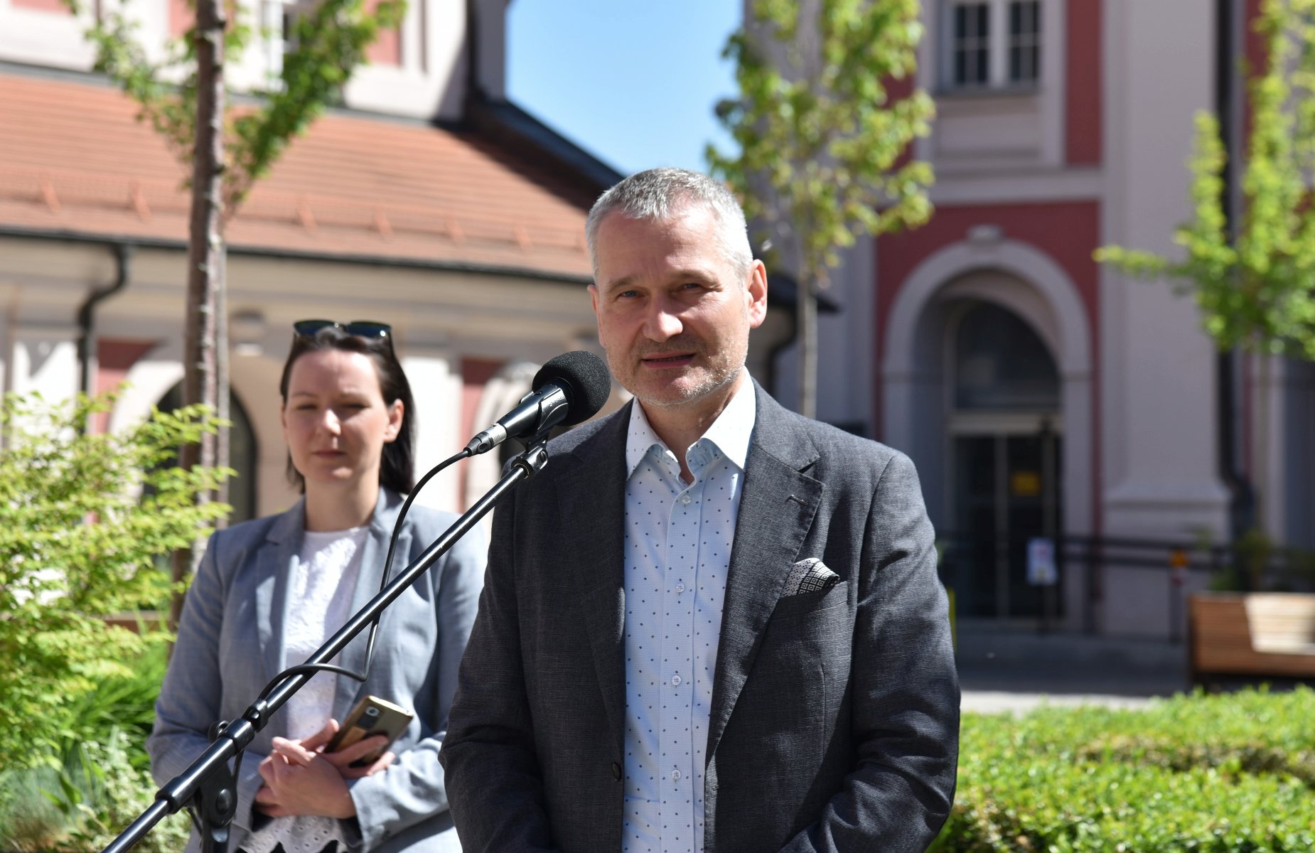 Na zdjęciu dwie osoby na dziedzińcu, mężczyzna przy mikrofonie, za nim kobieta - grafika rozmowy