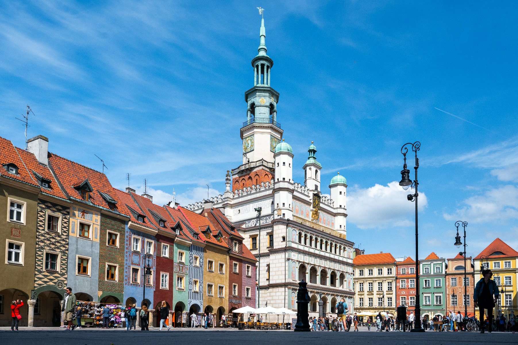 Zdjęcie przedstawia Stary Rynek w Poznaniu. W centralnej części kadru widać ratusz i domki budnicze, przed którymi przechadzają się mieszkańcy. - grafika rozmowy