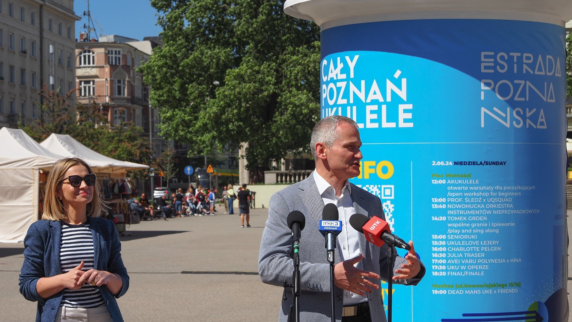 Na zdjęciu mężczyzna przy mikrofonie, obok stoi kobieta, w tle słup z plakatem festiwalu Cały Poznań ukulele