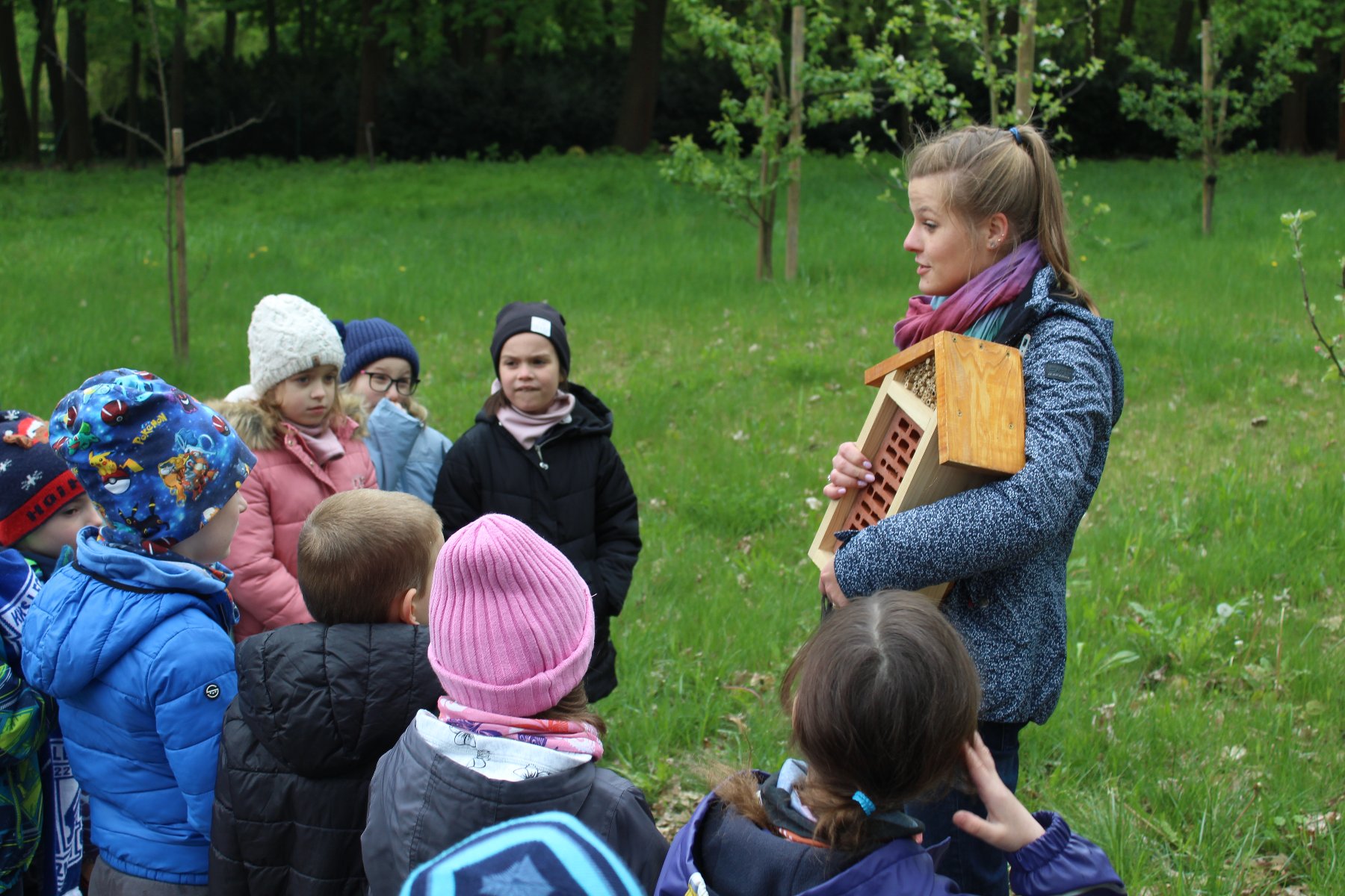 Zdjęcie przedstawia grupę dzieci i kobietę trzymającą domek dla owadów. - grafika rozmowy